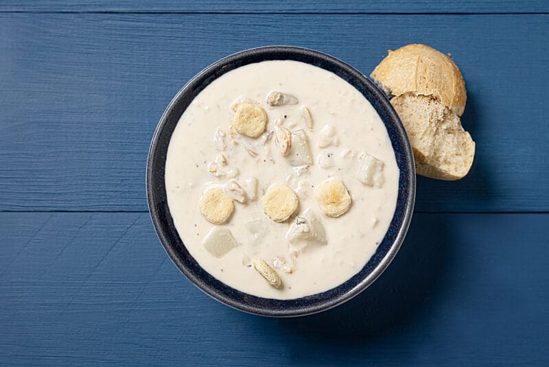 A bowl of creamy clam chowder garnished with oyster crackers is set on a blue wooden table. A partially broken bread roll sits to the side, ready to be enjoyed with the soup.