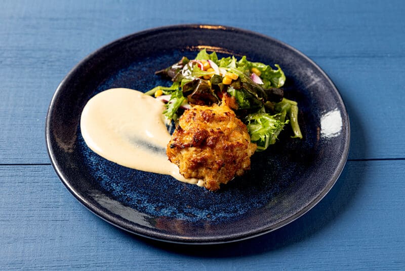 A dark blue plate containing a golden-brown crab cake with a dollop of creamy white sauce beside it, accompanied by a fresh mixed green salad with corn and small pieces of radish. The plate is set on a blue wooden table.