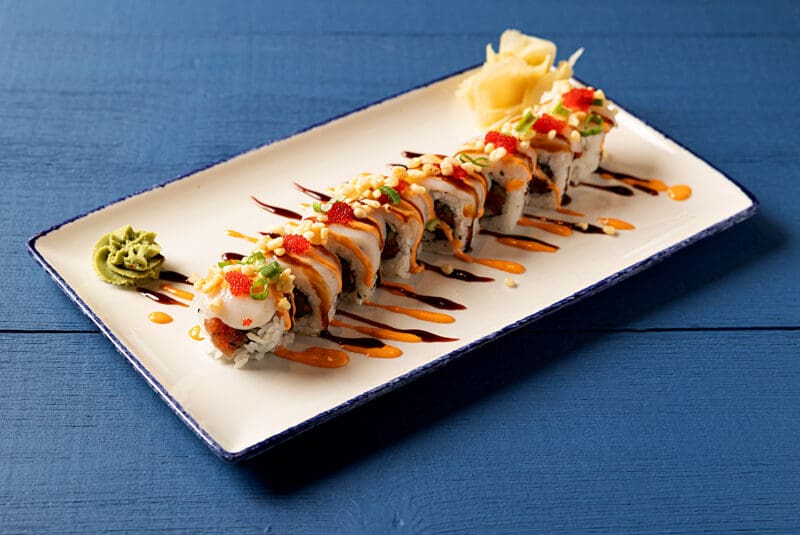 A rectangular white plate with blue edges holds a neatly arranged sushi roll topped with sauces, roe, and chopped green onions. A small mound of wasabi paste and a serving of pickled ginger are placed at either end of the plate on a blue wooden table.