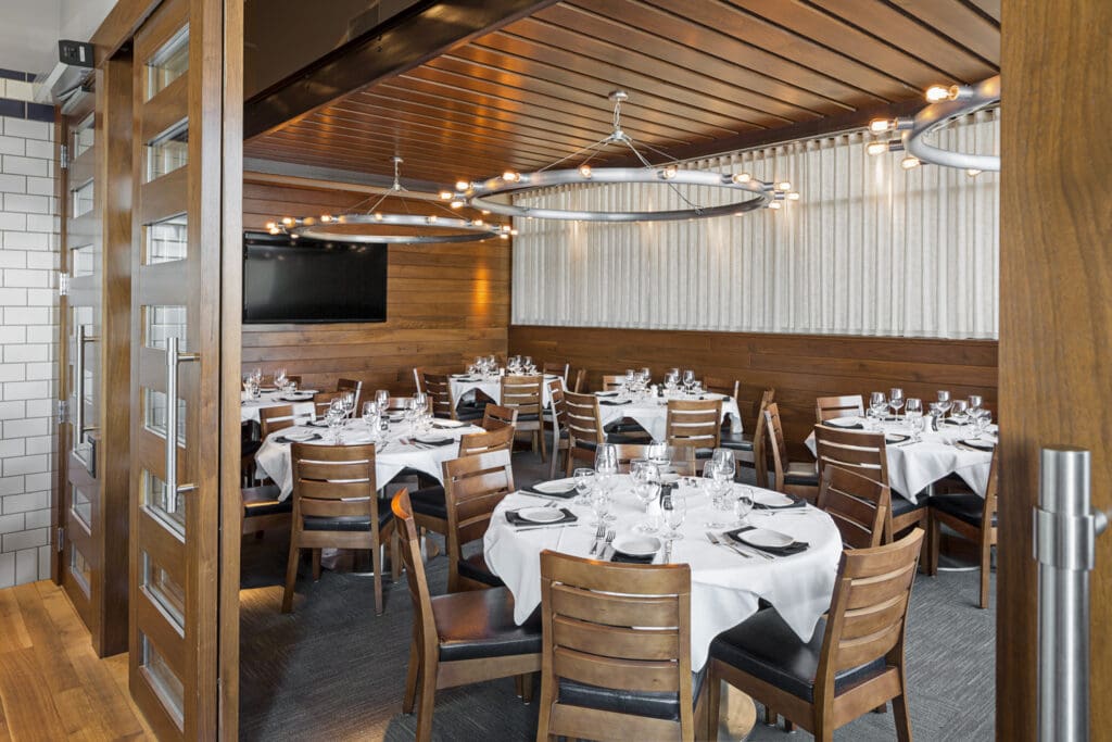 A small, elegantly decorated private dining room with round tables covered in white tablecloths. Wooden chairs surround the tables, each set with glassware, plates, and black napkins. Modern circular chandeliers hang from the wooden ceiling.