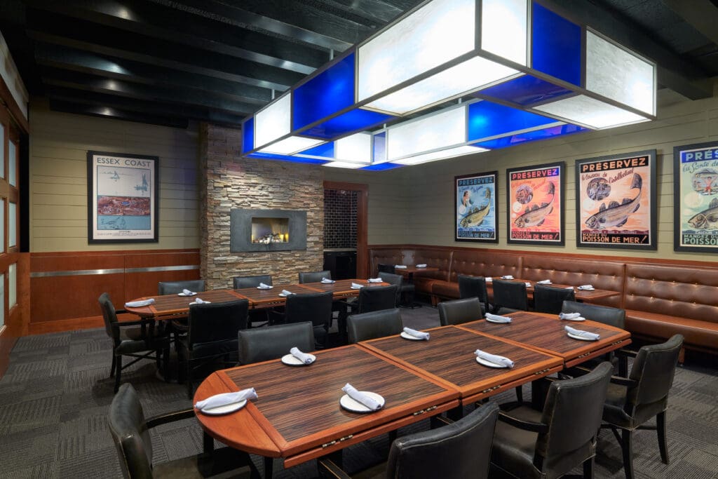 A modern dining room featuring a large wooden table set with white napkins, surrounded by black chairs. The walls display vintage posters, and a stone fireplace adds a cozy touch. The ceiling has a geometric overhead light fixture with blue accents.