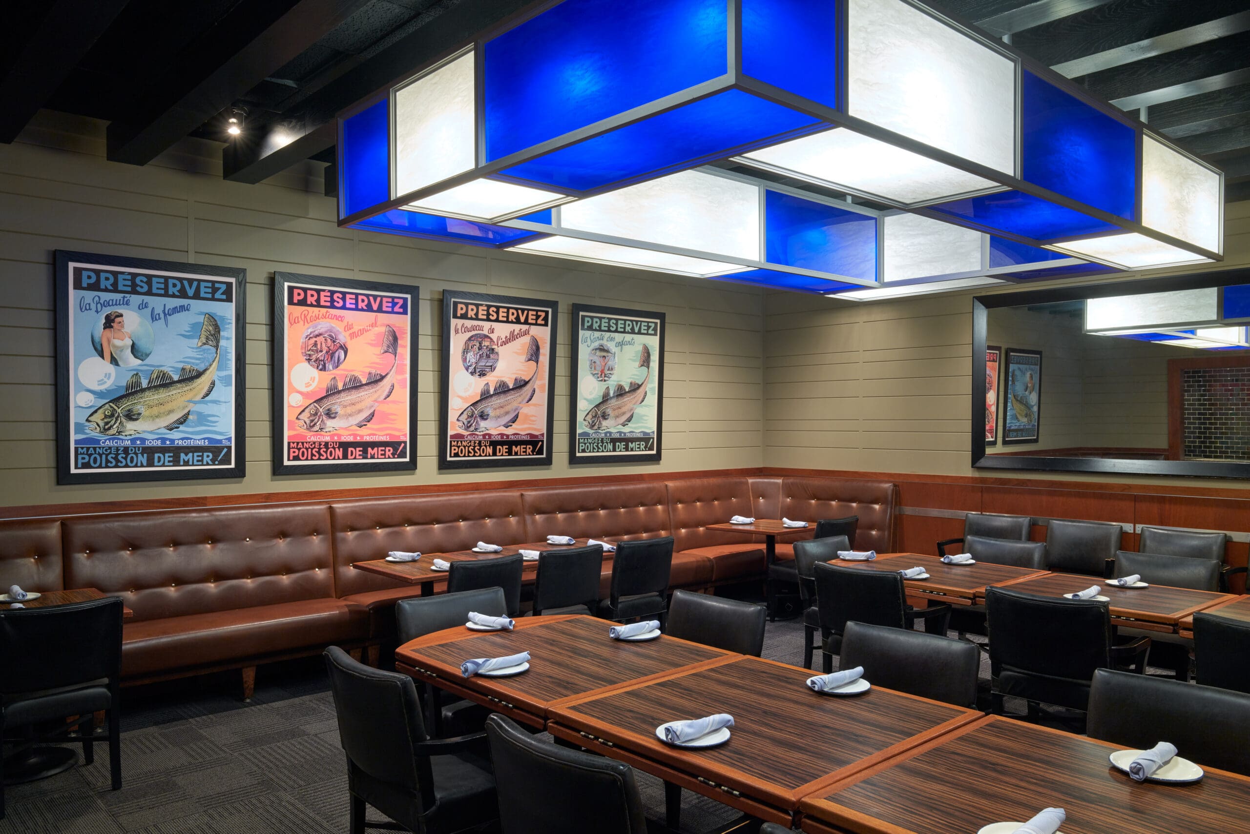 A restaurant interior features a long brown leather bench against the wall, with wooden tables and black chairs arranged neatly in front. Above the tables are modern blue and white hanging lights. The walls display vintage-style fish-themed posters.