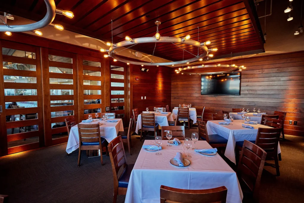 A cozy restaurant interior featuring several tables covered with white tablecloths and set with glassware and napkins. Modern, circular light fixtures hang from the wooden ceiling, and there's a flat-screen TV mounted on the wooden wall in the background.