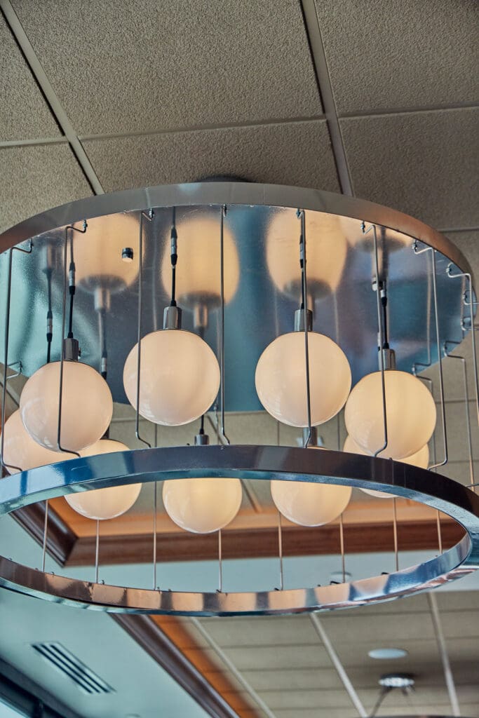 A modern light fixture with six spherical frosted glass bulbs is mounted on the ceiling. The fixture has a sleek metallic frame with two parallel circular bands, supported by vertical rods. The ceiling has a grid of square tiles, adding to the contemporary look.