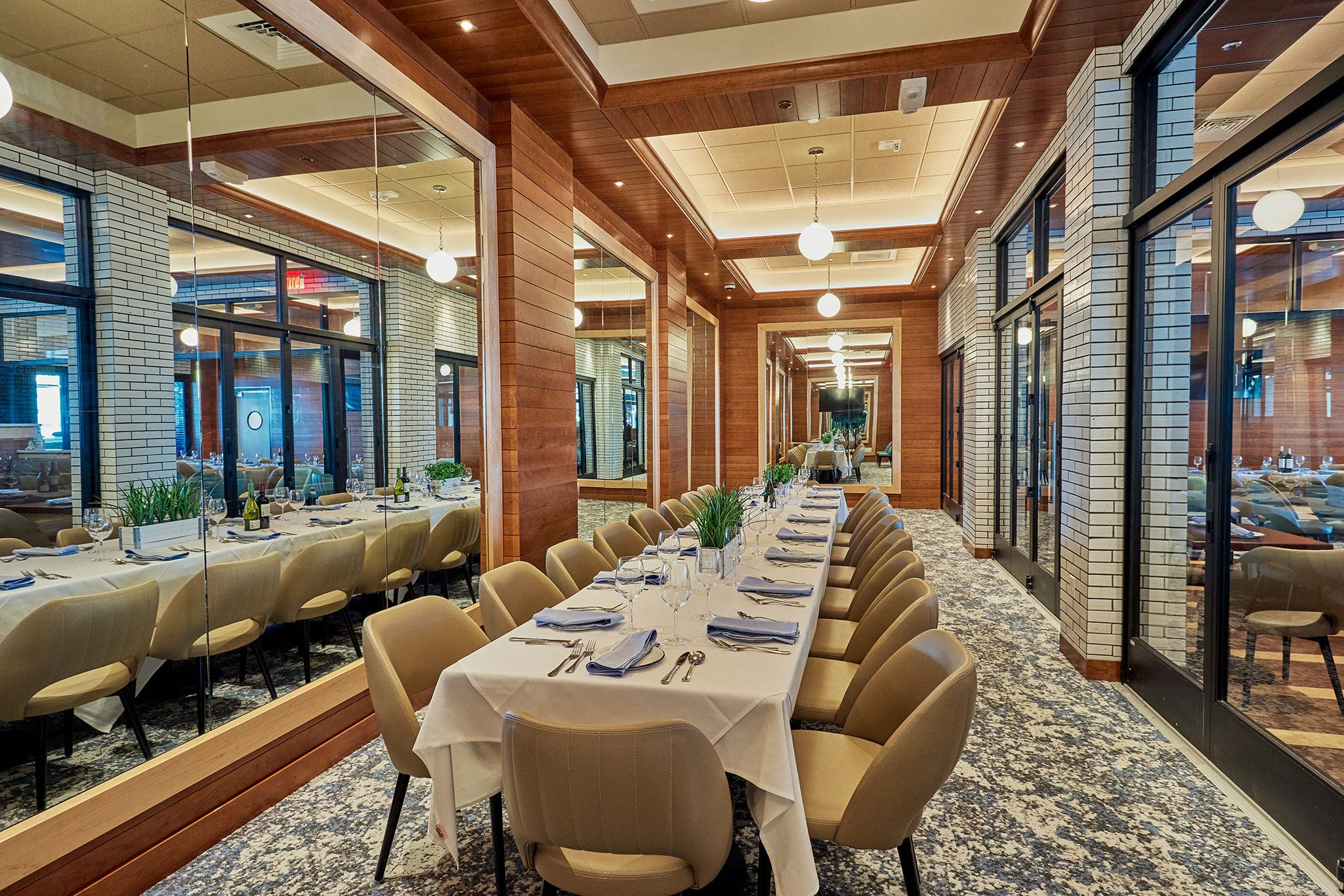 A long, elegantly set dining table is surrounded by beige chairs in a modern restaurant. The room features wooden accents, mirrored walls, and a tiled floor. The table is adorned with white tablecloths, silver cutlery, and green centerpieces. Spherical lights hang from the ceiling.