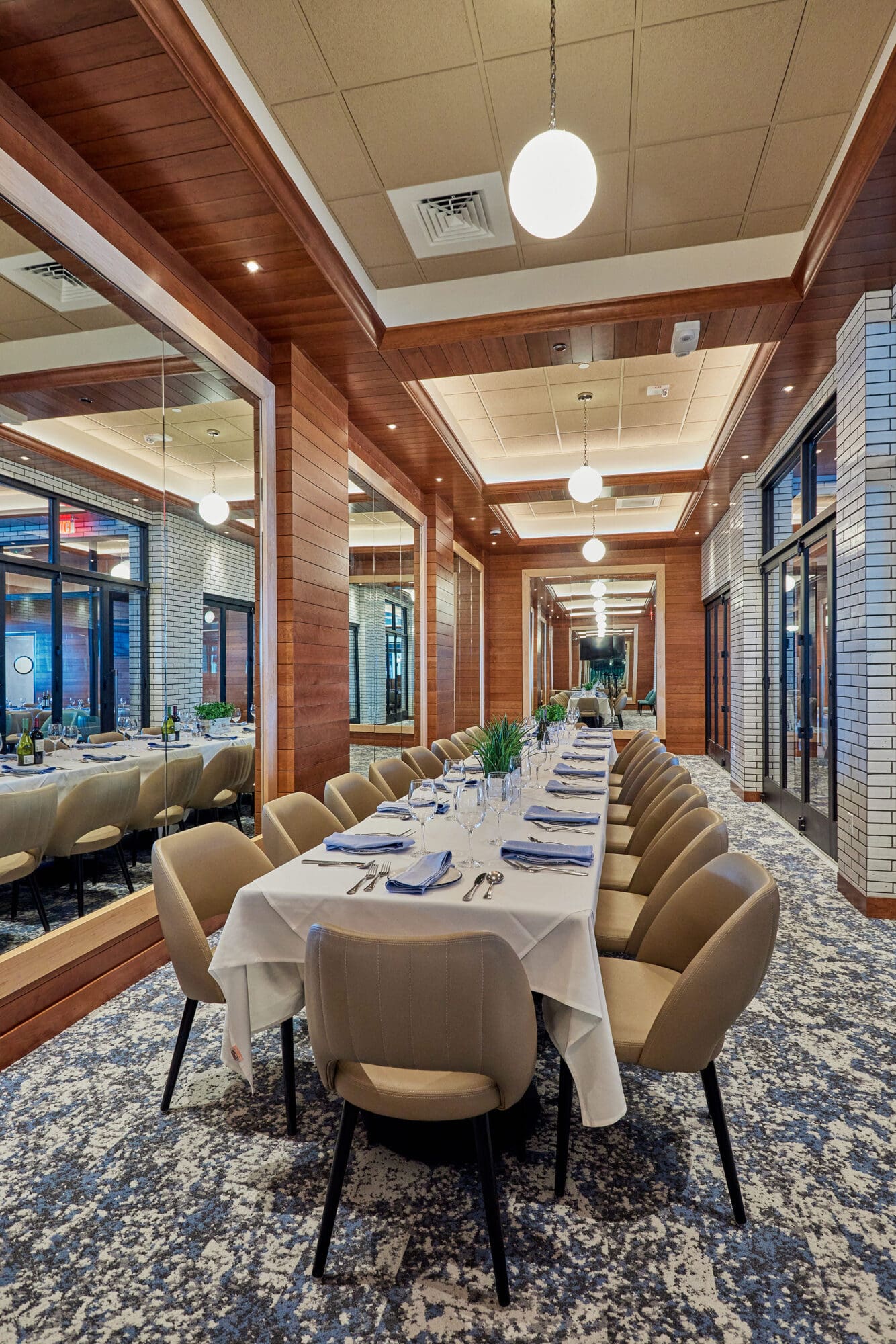 A modern, elegant dining room with a long table set for a meal in the center. The table is adorned with white tablecloths, neatly arranged plates, silverware, and glassware. Beige cushioned chairs surround the table. The room has wooden accents and mirrored walls.