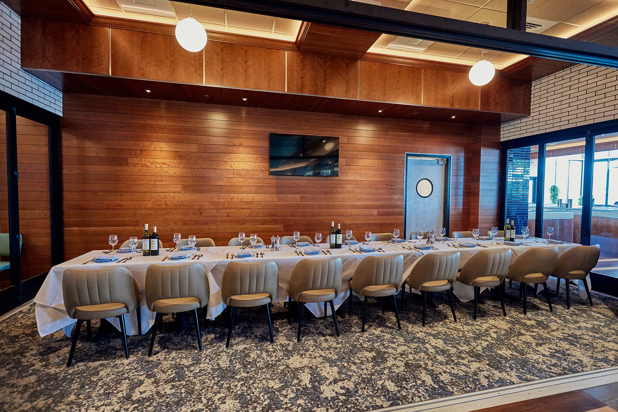 A long dining table set for twelve with white tablecloth, wine glasses, and cutlery is in a modern, elegantly decorated room. The walls feature wooden paneling and a flat-screen TV. Ceiling lights illuminate the space, and large windows allow natural light to enter.