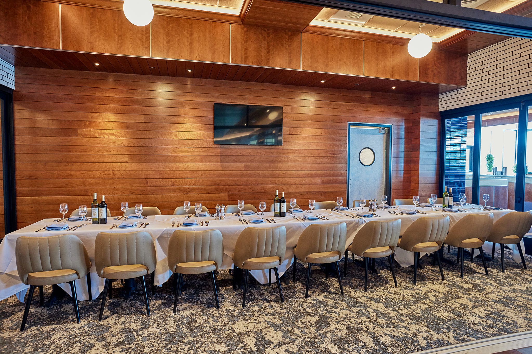 A long dining table set for a formal meal in an elegant room with wood-paneled walls. The table is adorned with white tablecloths, wine bottles, glasses, plates, and cutlery. Beige cushioned chairs surround the table, and a flat-screen TV is mounted on the wall.