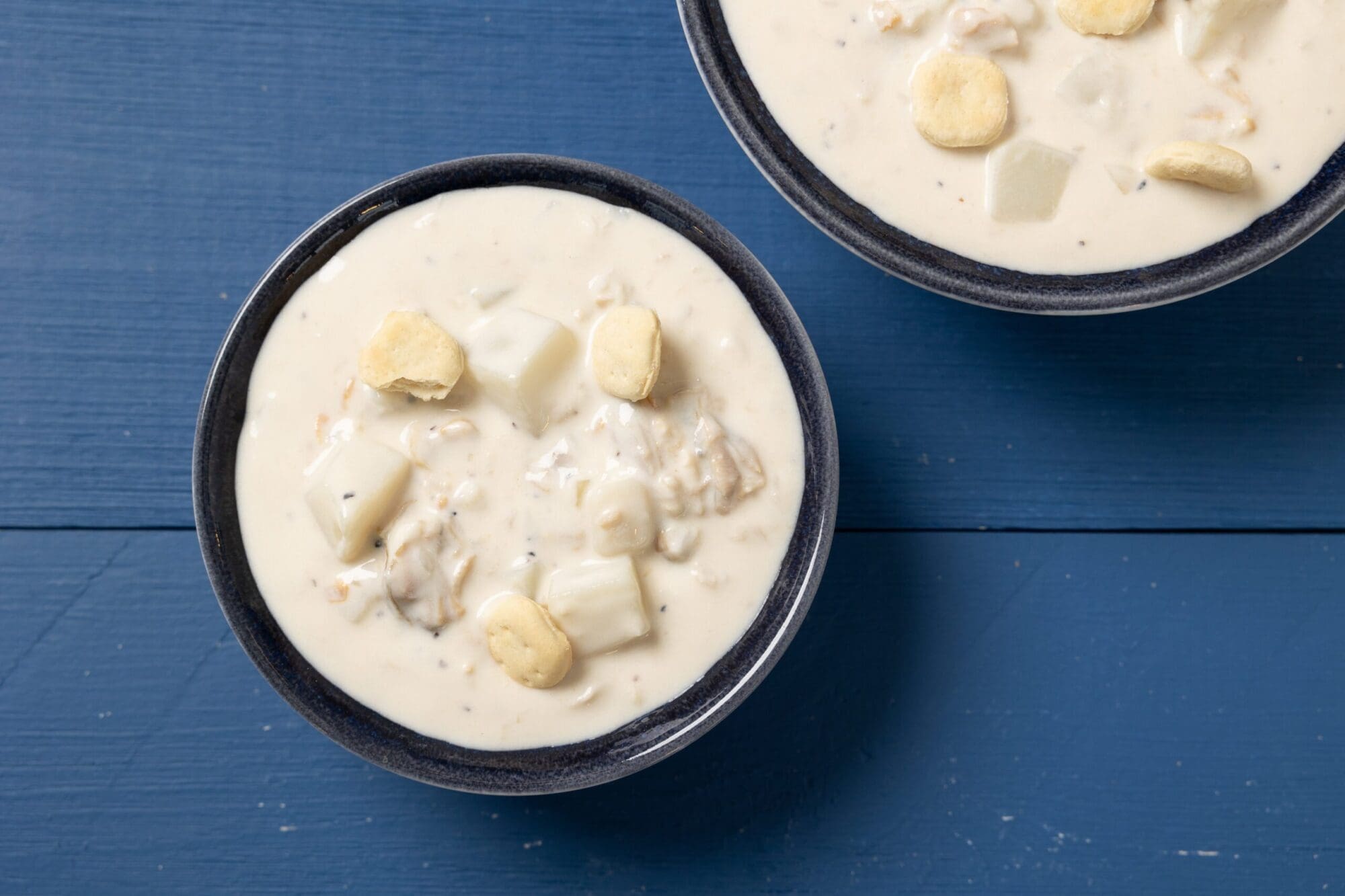 Two bowls of creamy clam chowder topped with oyster crackers. The chowder has chunks of potatoes and clams, and it is served in dark-colored bowls placed on a blue wooden surface.