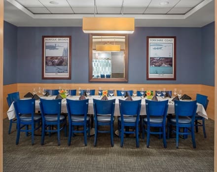 A long dining table with blue chairs arranged neatly around it is set up in a formal dining room. The table is adorned with glasses, plates, and vibrant floral centerpieces. The walls are decorated with framed posters and a large mirror.