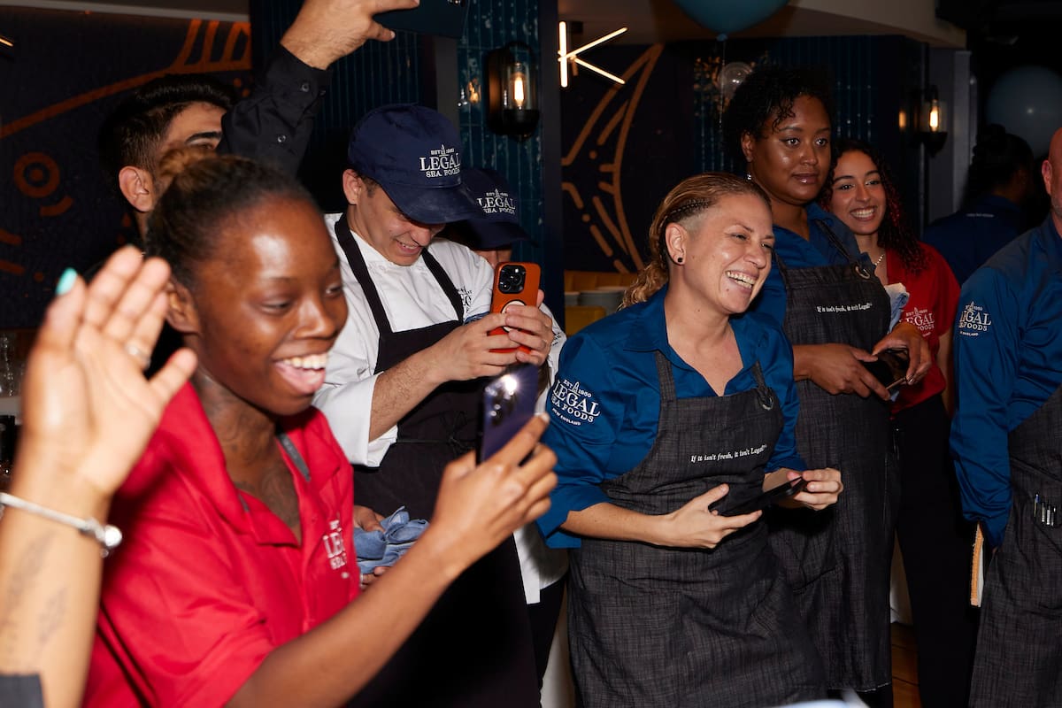 A group of people are standing together indoors, smiling and looking at their phones. Some wear uniforms and aprons. They appear to be in a celebratory mood, possibly at a restaurant or event venue. Blue and red shirts are visible among the group.