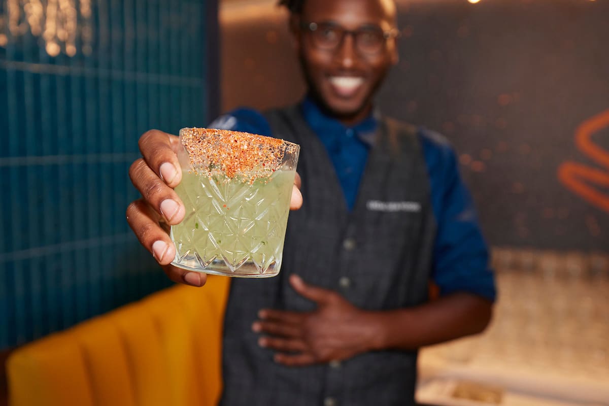 A person in a blue shirt, tie, and gray vest, smiling and holding up a glass filled with a green beverage. The glass has a chili-salt rim. The background features a teal-tiled wall and a yellow booth.