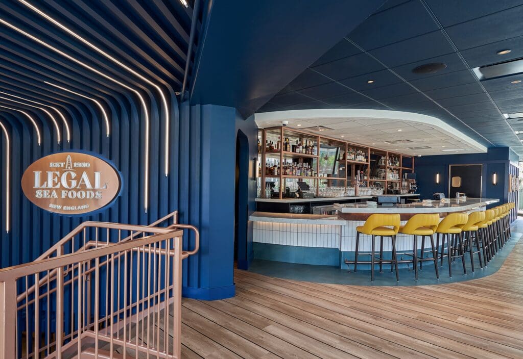 A modern restaurant interior showcasing a bar area with a curved counter and yellow bar stools. The walls and ceiling are dark blue, complemented by wood flooring. A sign on the left reads 