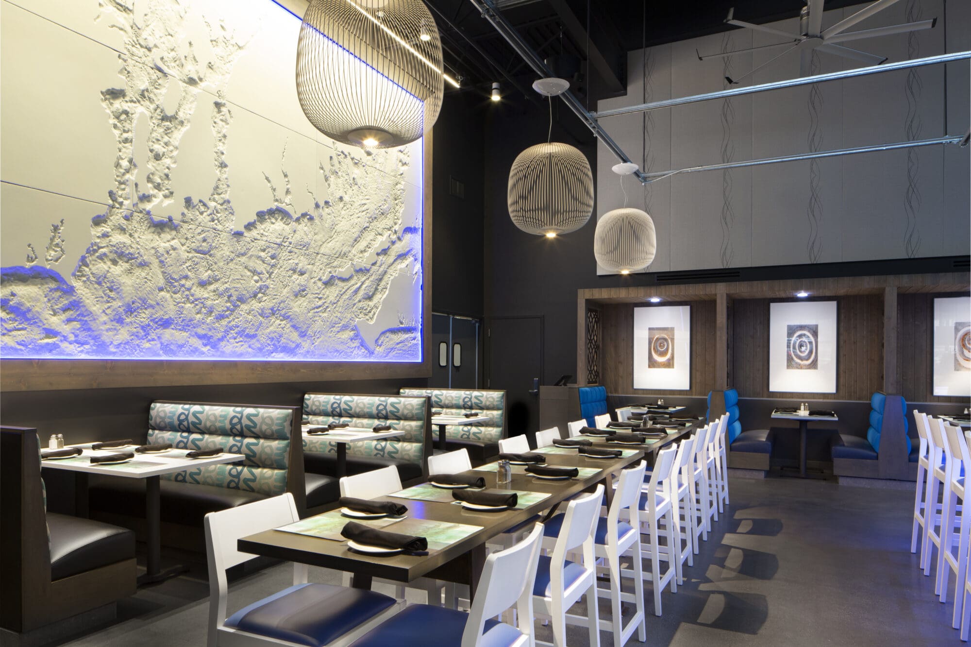 A modern restaurant with a mix of booth and chair seating. The walls are decorated with textured art and framed pictures. Black tables are set with dishes and napkins. Several pendant lights hang from the ceiling, which features exposed beams and ducts.