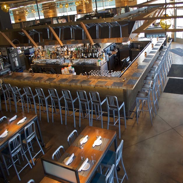 The image shows the interior of a modern bar and dining area. The bar features a long counter with numerous metal barstools arranged neatly. Behind the bar, a variety of alcoholic beverages are displayed. Nearby, wooden dining tables and chairs are set for customers.