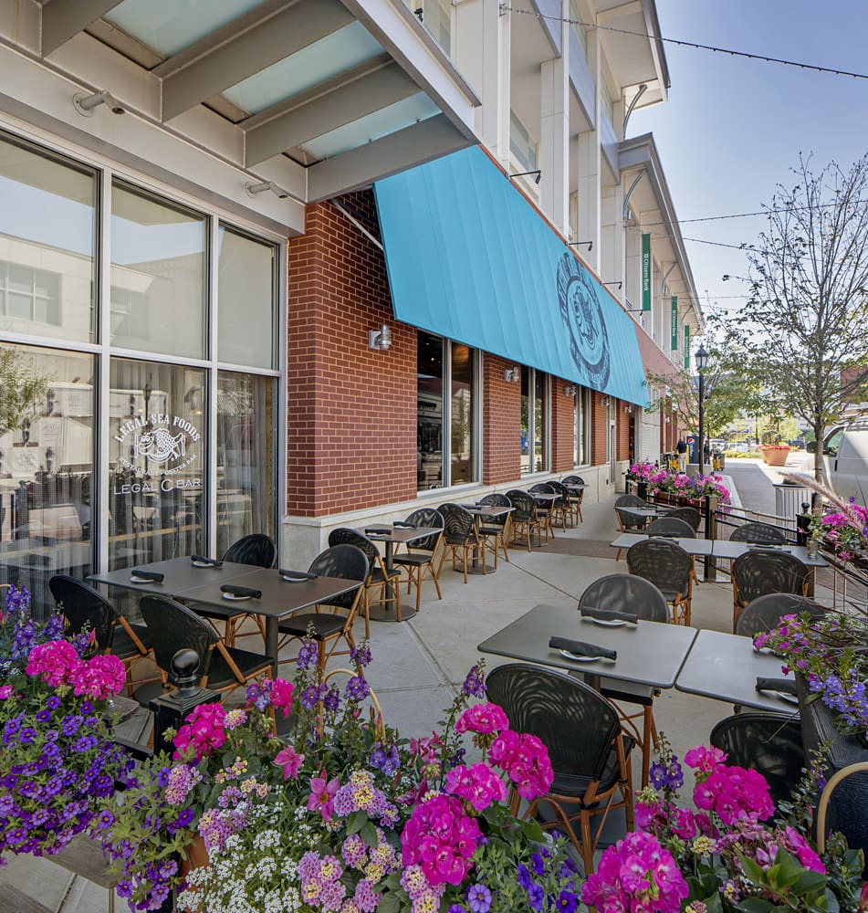 A charming outdoor seating area of a restaurant with several tables and chairs. The space is adorned with vibrant pink and purple flowers. The restaurant features a red brick exterior and a large blue awning with a logo. The scene is well-lit and inviting.