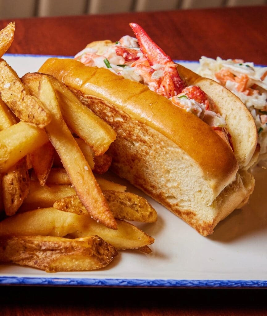 A plate features a lobster roll with chunks of lobster meat inside a toasted bun, accompanied by a pile of golden-brown French fries. The dish is served on a white plate with blue trim, set on a wooden table.