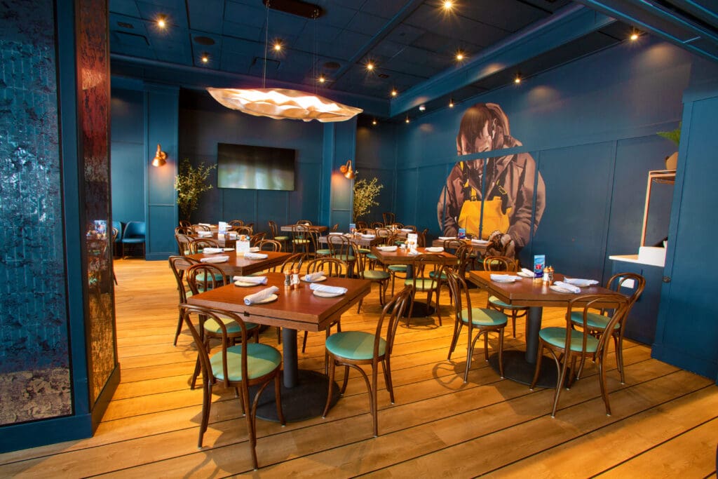 A modern, stylish restaurant interior with wooden floors and tables, surrounded by dark blue walls. The tables are set with white napkins and cutlery, and there is a large mural of a person on one wall. A unique light fixture hangs from the ceiling.