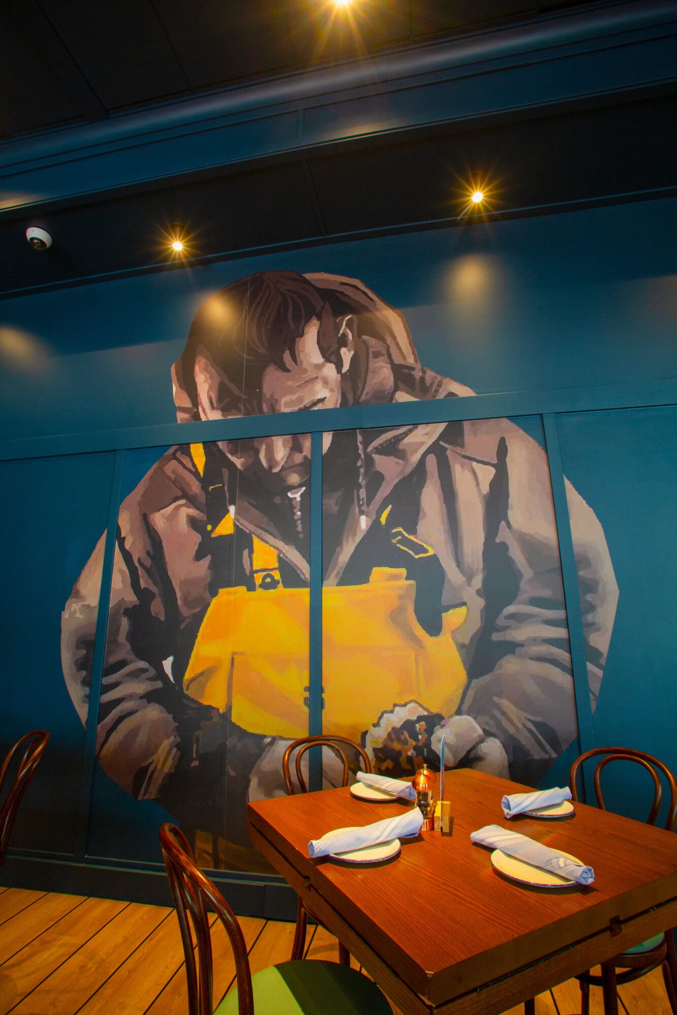 A dimly lit restaurant features a large mural on a teal wall, depicting a weathered fisher in a yellow raincoat. In the foreground, a wooden table is set with white napkins, plates, and cutlery, surrounded by wooden chairs. The atmosphere is cozy and intimate.