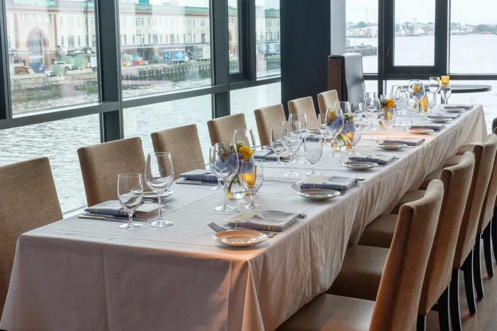 A long dining table is elegantly set with white tablecloth, neatly arranged wine glasses, silverware, and small floral centerpieces. Brown upholstered chairs surround the table. Large windows offer a view of a waterfront with buildings and boats in the background.