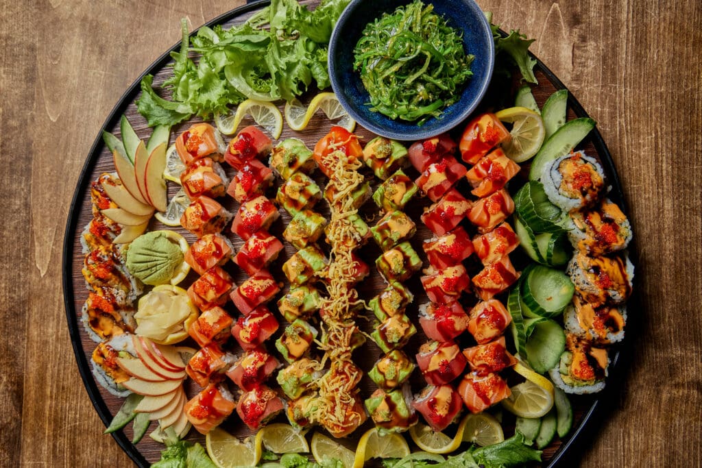 A vibrant platter of assorted sushi rolls, garnished with sauces and toppings, arranged in rows on a wooden surface. The platter also includes slices of lemon, apple, lime, avocado, wasabi, and pickled ginger, with a bowl of seaweed salad in the center.