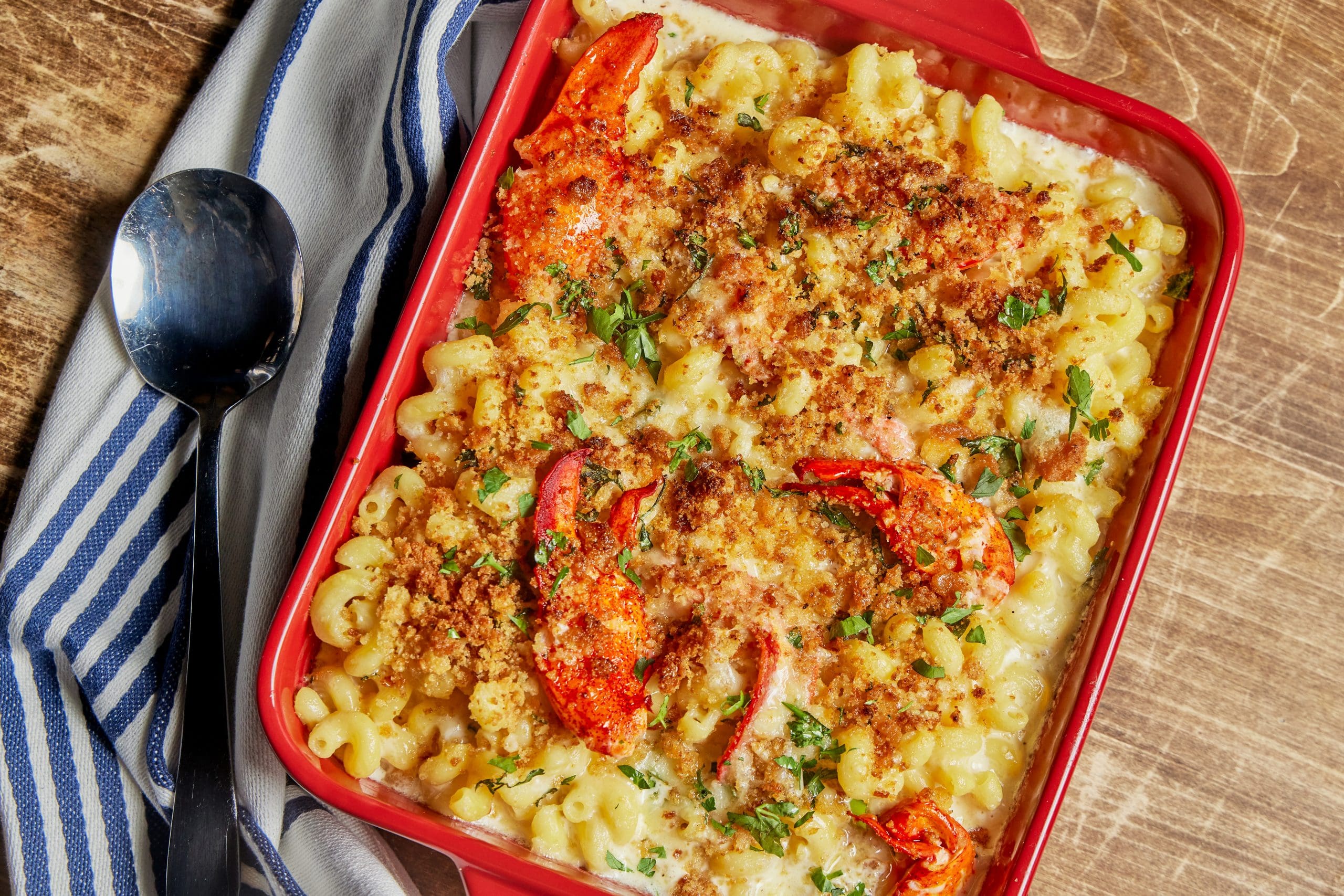 A red baking dish filled with creamy macaroni and cheese topped with a golden breadcrumb crust and garnished with herbs and roasted red pepper slices. A striped blue and white kitchen towel and a spoon are placed next to the dish on a wooden surface.