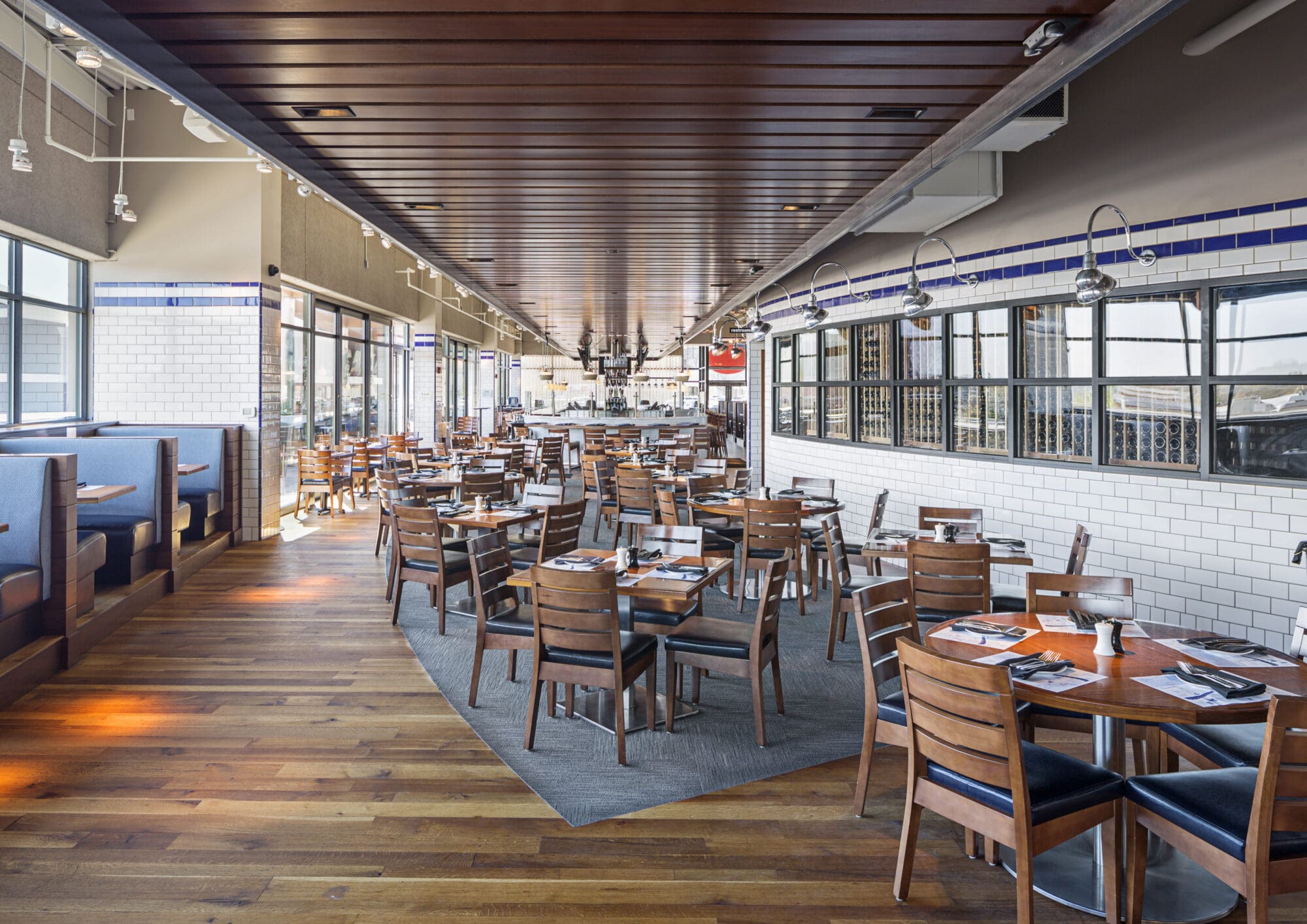 A spacious, well-lit restaurant interior with wooden flooring and ceiling. The seating area features wooden tables and chairs, as well as blue upholstered booths along the windows. Large windows line one side, allowing natural light to flood the space.