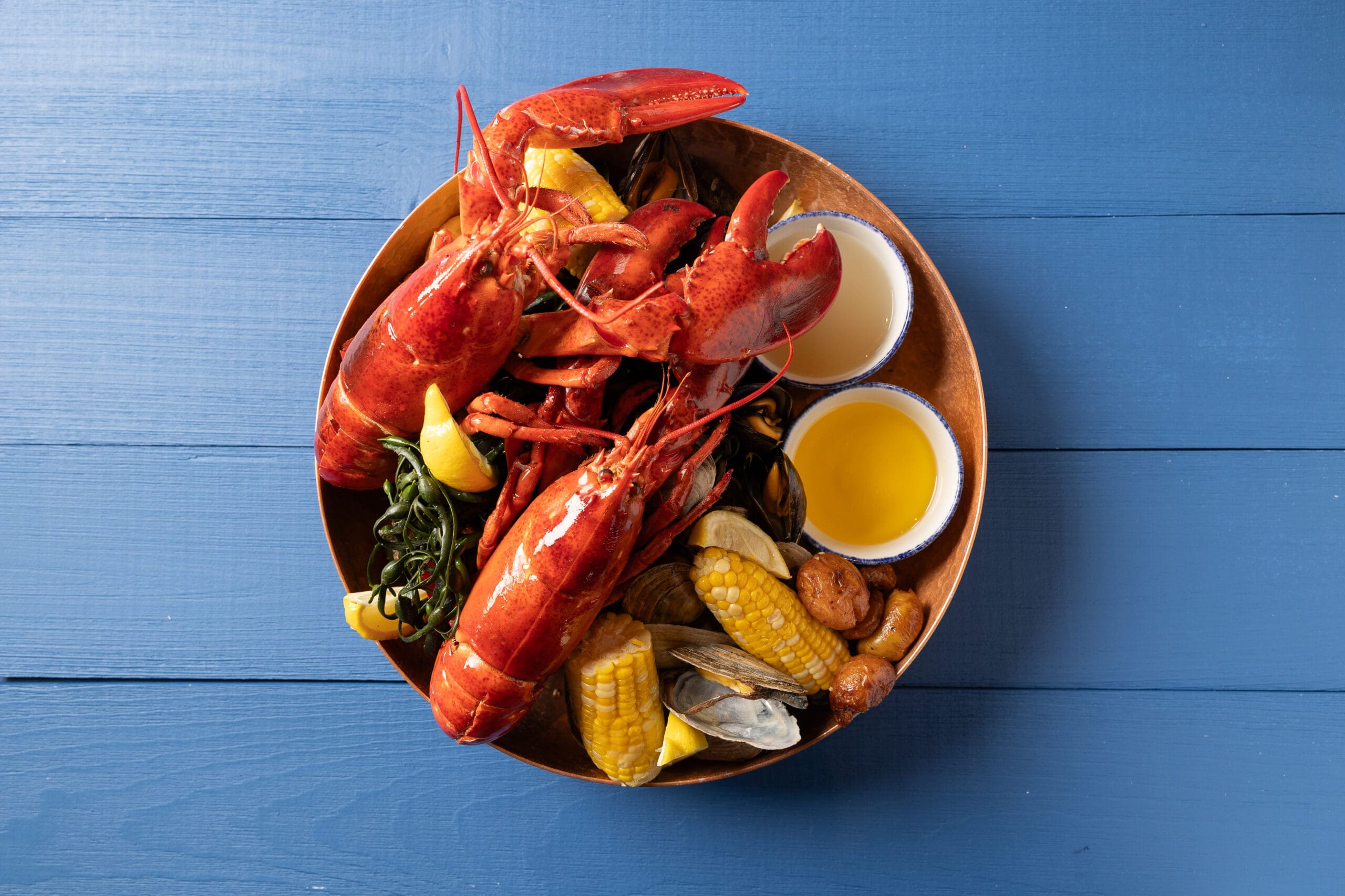 A wooden bowl filled with a seafood feast; two whole lobsters, corn on the cob, clams, mussels, sausage pieces, seaweed, and lemon wedges. Two small bowls of melted butter are also included. All items are arranged on a blue wooden surface.