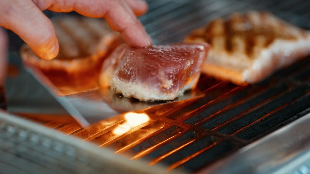 Close-up of a hand using a spatula to grill pieces of meat over an open flame on a barbecue grill. The meat has visible grill marks, indicating it is being cooked. The image captures the process of grilling food outdoors.