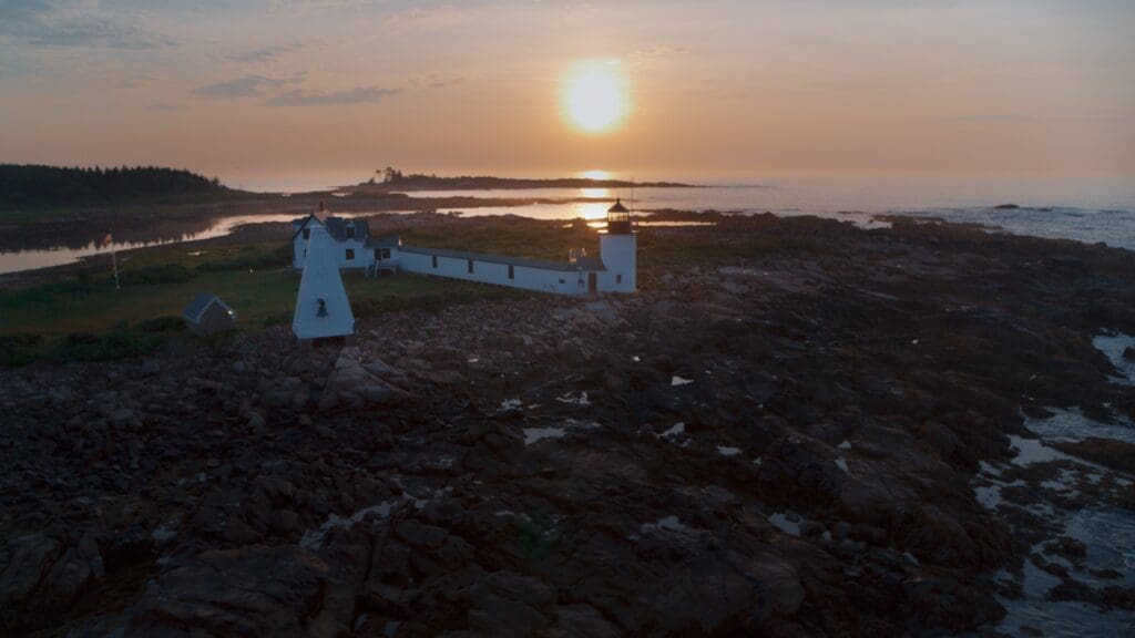 An aerial view of a coastal landscape at sunset shows a lighthouse and connected buildings on rocky terrain. The lighthouse stands near the shore, with the sun setting over the water, casting a warm glow on the scene. The coastline and distant trees are visible.