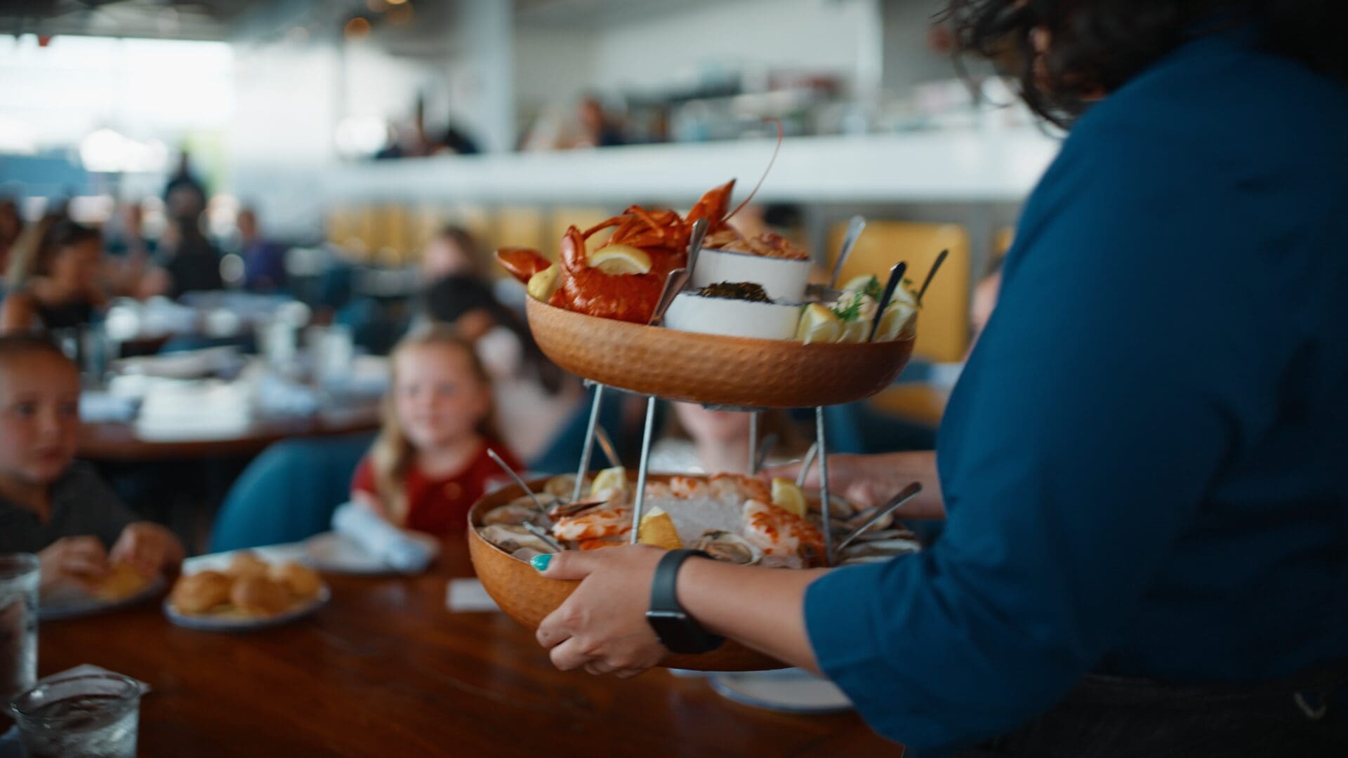 A restaurant server carries a two-tiered platter filled with seafood dishes, including lobsters and oysters, towards a table where children are seated, eagerly awaiting their meal. The dining area features a modern, casual ambiance with other diners in the background.