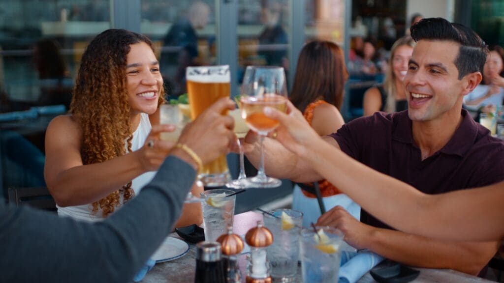 A group of people are seated at an outdoor restaurant table, smiling and raising their glasses in a toast. The table is set with drinks and tableware. The background shows other diners and a glass window reflecting the outdoor scene.