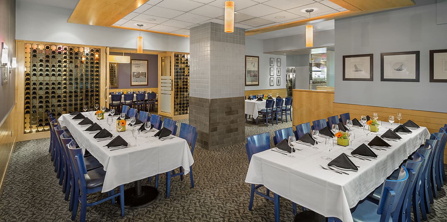 A restaurant dining area is set up with white tablecloths and black napkins on the tables. Blue chairs surround the tables. The walls have framed pictures, and there is a wine rack in an alcove. Pendant lights hang from the ceiling, illuminating the space.