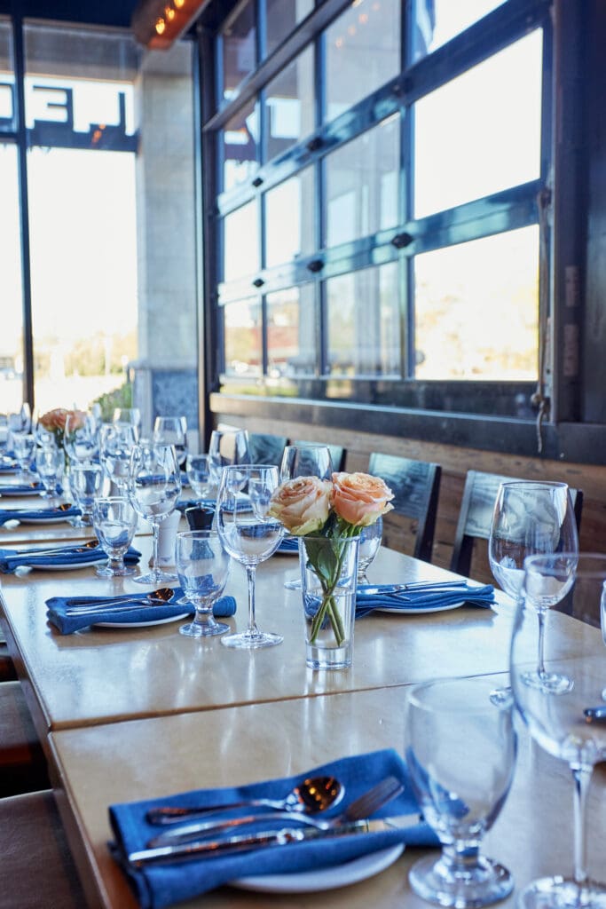 A long dining table set for a meal with blue napkins, empty wine glasses, and water glasses. Small vases with peach-colored roses serve as centerpieces. The table is positioned near large windows, allowing natural light to illuminate the setup.