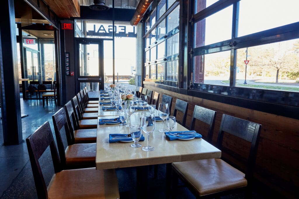 A long dining table set with wine glasses, blue napkins, and small floral centerpieces is arranged in an airy, modern restaurant with large windows and wooden accents. Natural light illuminates the space through the open door and wall of windows.