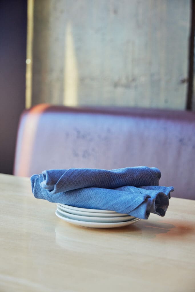 A neatly folded blue cloth napkin rests on a stack of white plates on a light-colored wooden table. The background includes part of a wooden booth with a purple cushion, softly lit by natural light streaming in.