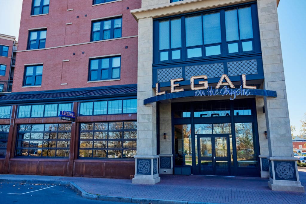 A brick building with a sign reading “Legal on the Mystic” above the entrance, indicating it is likely a restaurant. The entrance features large windows and stone columns. There are small trees reflected in the windows and a clear, sunny sky overhead.