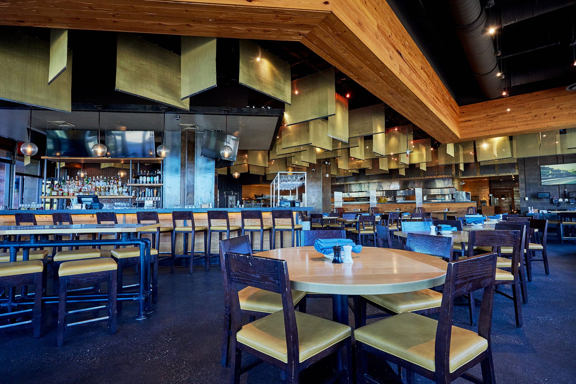 A modern restaurant interior with wooden tables and chairs, a long bar with barstools, hanging light fixtures, and an open kitchen in the background. The ceiling features wood and metallic panels. The space is well-lit and has a contemporary design.