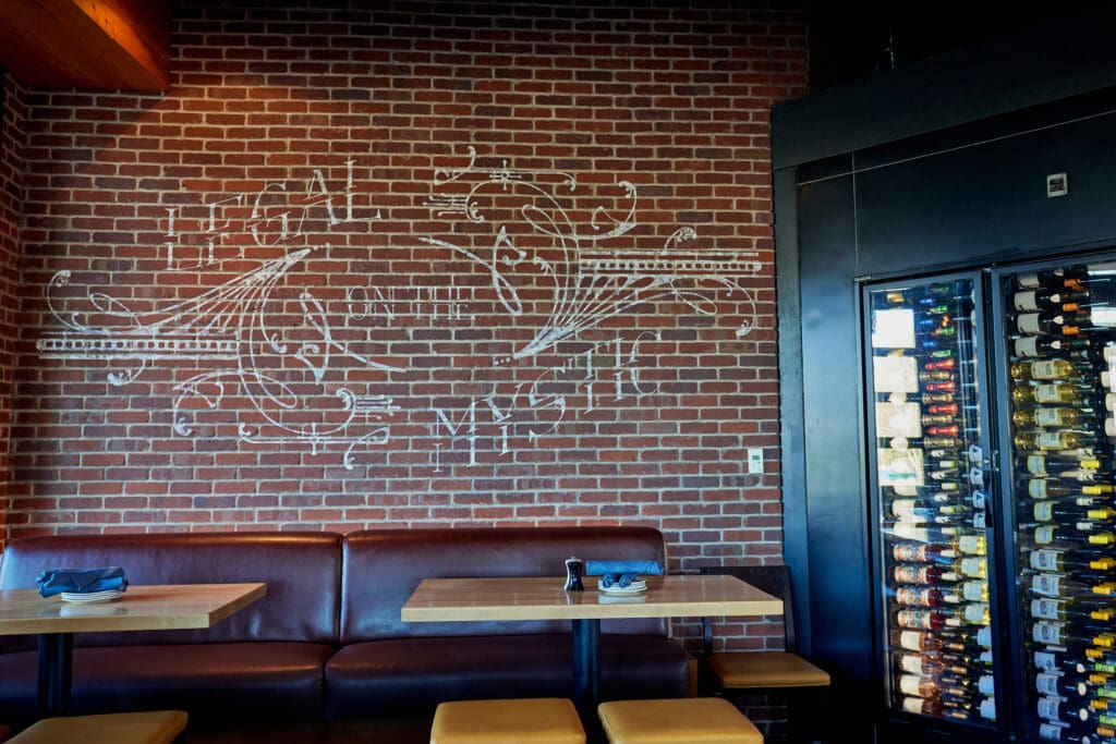 A restaurant interior with exposed brick walls, a leather bench, wooden tables, and stools. A chalkboard with 