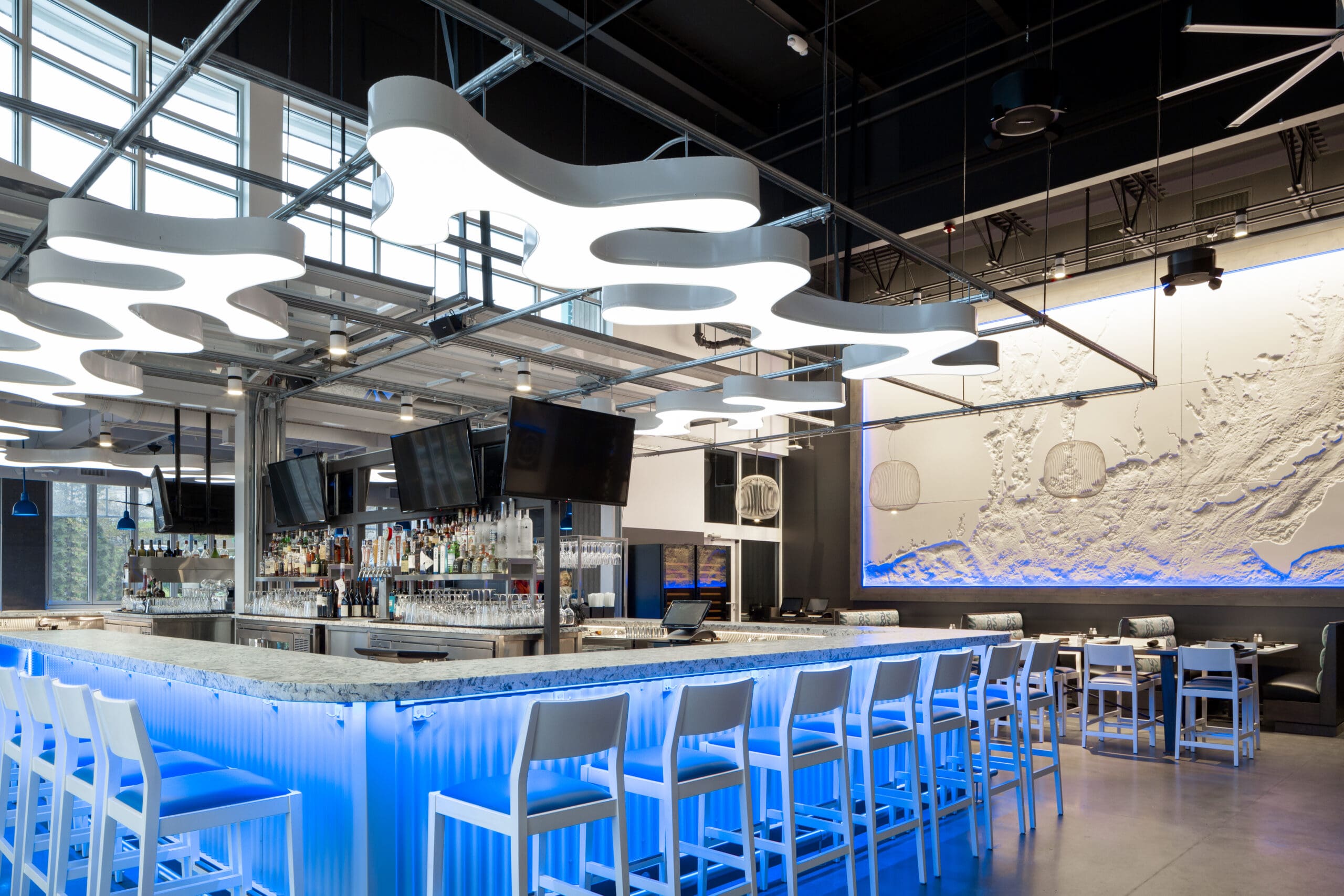 A modern bar with a futuristic vibe, featuring a white marble countertop and steel bar stools with blue cushions. Unique cloud-shaped ceiling lights and blue LED accent lighting create an ambient atmosphere. The back wall displays a textured map illuminated in blue.