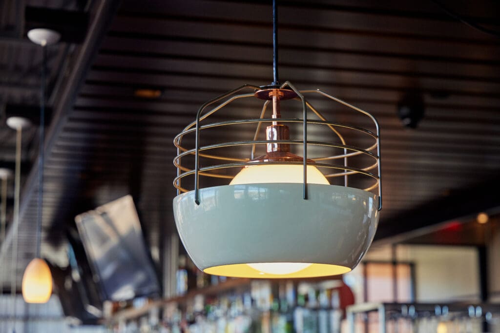 A modern pendant light with a white bowl-shaped shade and thin metal cage hangs from a dark ceiling. The background shows other pendant lights and TV screens, and the interior has a contemporary look with a bar area visible.