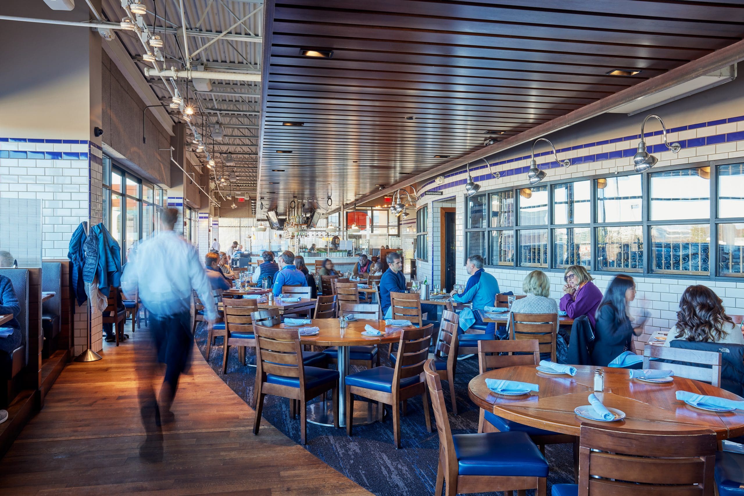 A modern, busy restaurant with wooden tables and chairs. Diners are seated, enjoying their meals, and waitstaff are moving between the tables. The interior features a sleek design with white tile walls, large windows, and a partially visible open kitchen area.