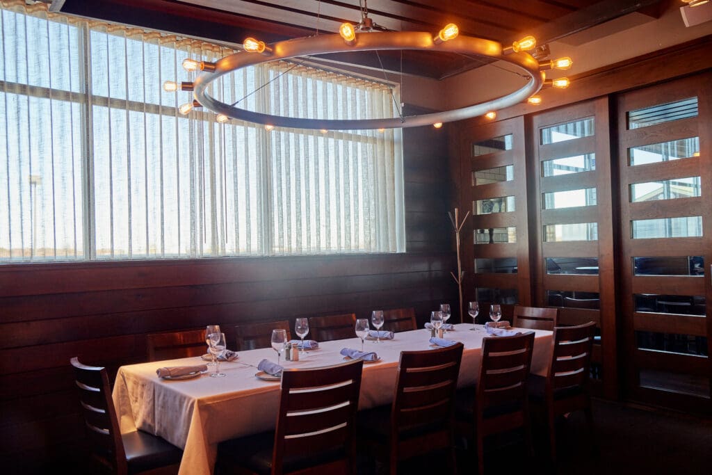 A rectangular dining table set for eight with white tablecloth and formal table settings is centered in a room with wooden paneling. An oval-shaped chandelier with warm lights hangs above. Large windows with vertical blinds allow natural light to hit the space.