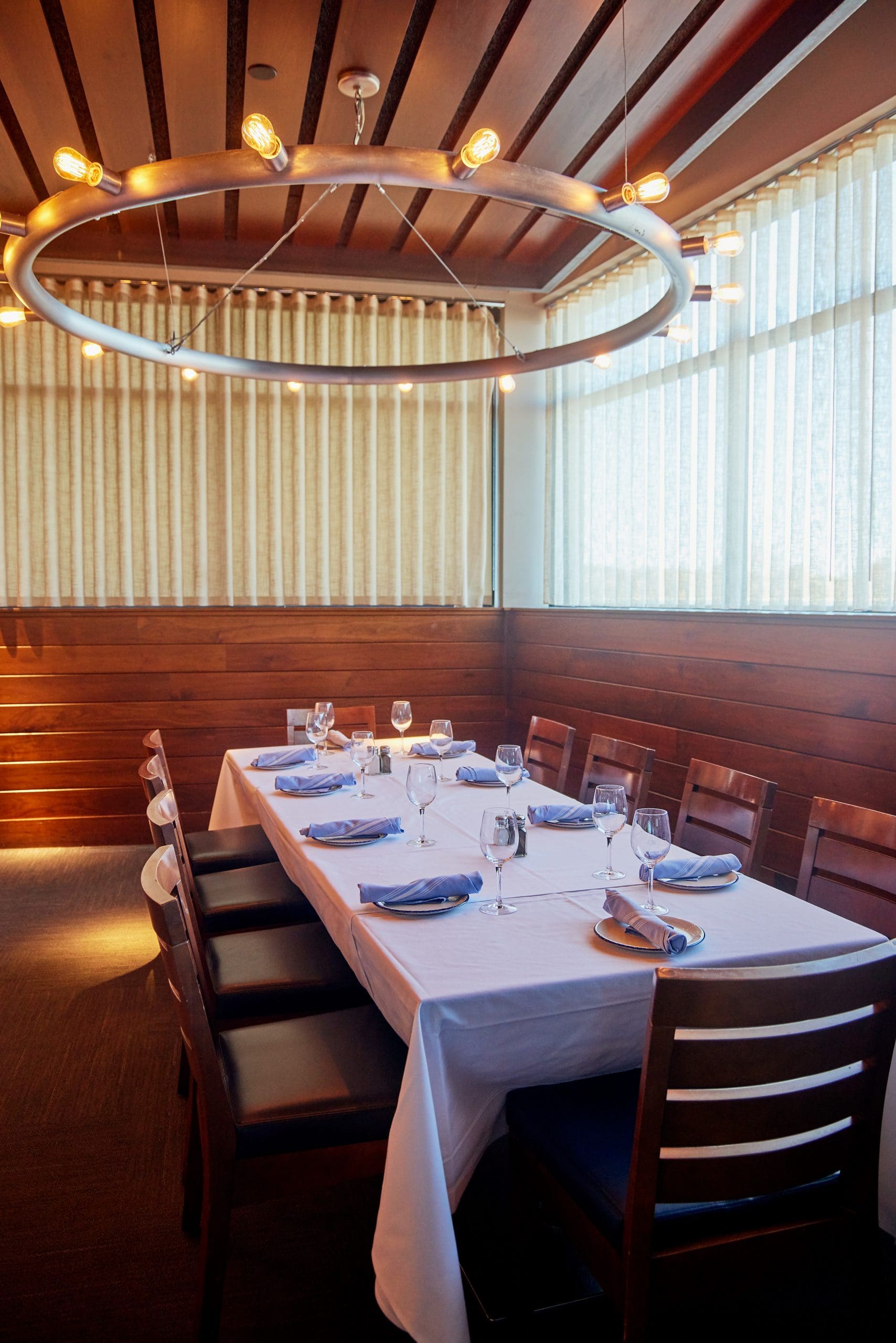 A well-lit private dining room featuring a rectangular table covered with a white tablecloth, set for eight people. The table is adorned with neatly folded blue napkins, wine glasses, and cutlery. Wooden chairs surround the table, and a modern circular light fixture hangs above.