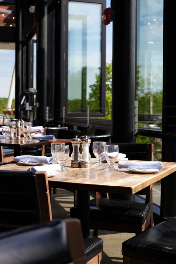 A sunny day in a restaurant with large windows. The wooden table is set with napkins, drinking glasses, and condiments. The table's surroundings include chairs and a scenic view of greenery outside the windows.