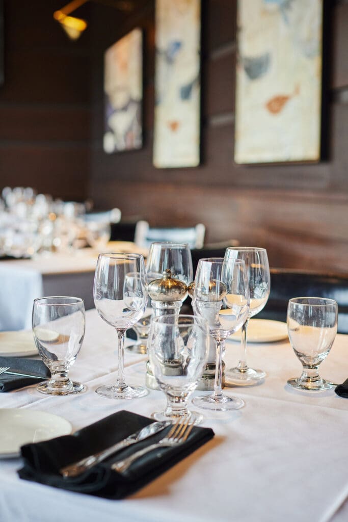 Table set for a fine dining experience, featuring multiple wine glasses, folded napkins with cutlery, a pepper grinder, and plates on a white tablecloth. Elegant paintings hang on the wooden-paneled wall in the background.