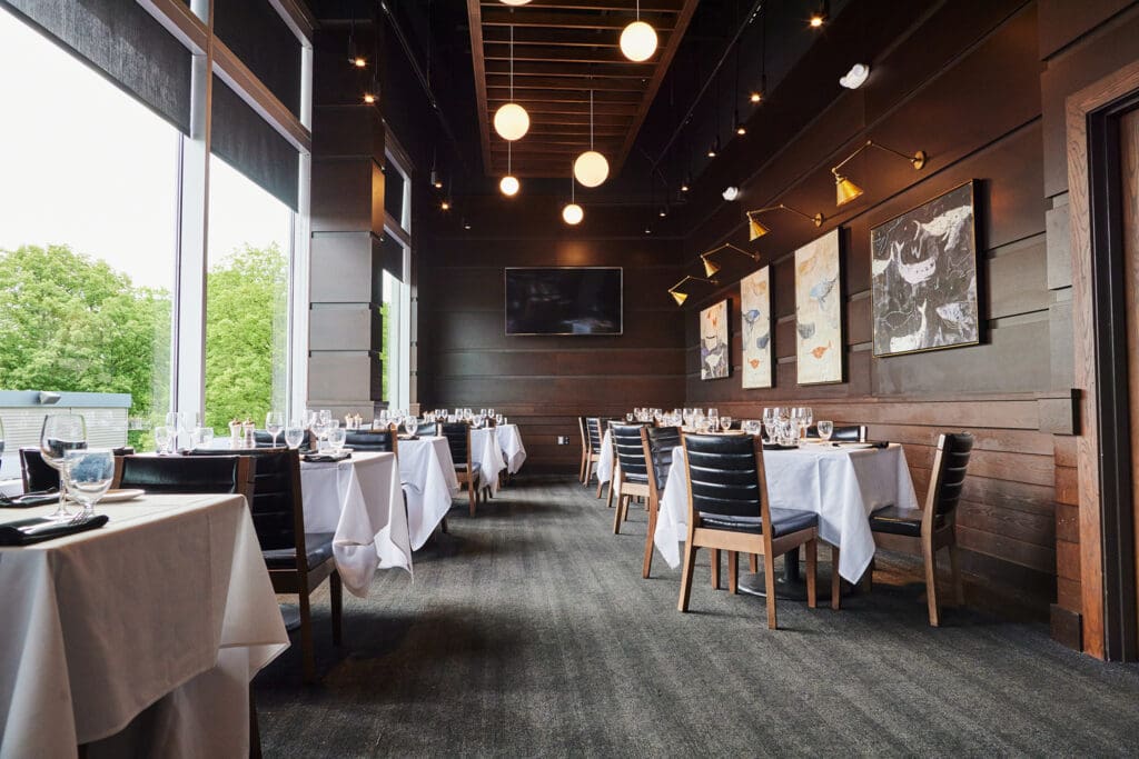 A sophisticated restaurant interior featuring wooden furniture, white tablecloths, and a serene ambiance. Modern art adorns the dark wooden walls, and round lights hang from the ceiling. Large windows offer natural light and views of greenery outside.