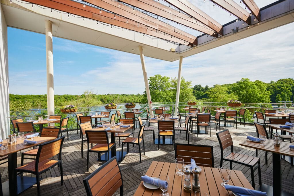 An outdoor restaurant patio with wooden tables and chairs, set with plates, glasses, and silverware. Overhead, a partial wooden pergola provides shade. The setting overlooks a lush green landscape with a view of a river or lake under a clear sky.