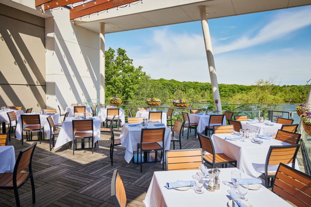 An outdoor restaurant seating area with several tables covered with white tablecloths and surrounded by wooden chairs. The tables are set with glassware and cutlery. The terrace overlooks a lush, green landscape under a clear blue sky.