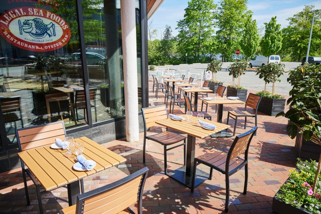 Outdoor dining area of Legal Sea Foods restaurant featuring multiple wooden tables with silverware, napkins, and wine glasses set up. The area is adjacent to the restaurant's glass windows, surrounded by greenery, and has a sunny, inviting atmosphere.